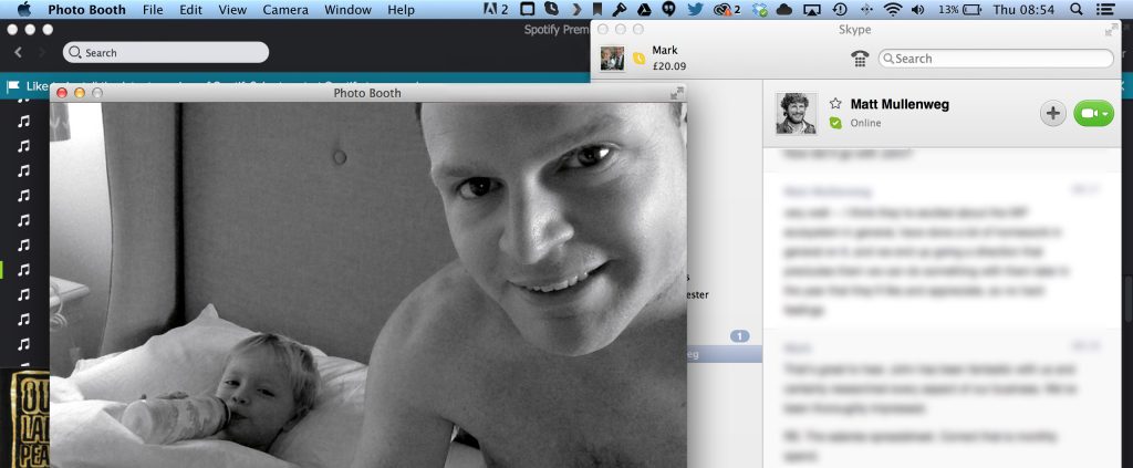 A black and white photo of a man smiling at the camera, holding a child in bed, with a computer screen displaying a Skype conversation and a Spotify interface in the background.